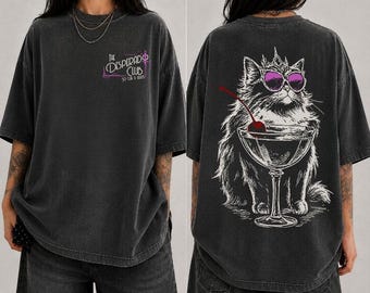 May include: A charcoal gray oversized t-shirt featuring a small graphic on the front that reads The Desperado Club. The back displays a large illustration of a fluffy cat wearing a crown and purple sunglasses, sitting beside a cocktail glass.