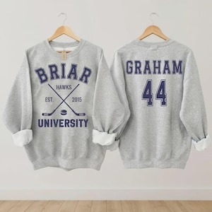 May include: A heather gray sweatshirt with the text "BRIAR UNIVERSITY HAWKS" in navy blue, along with crossed hockey sticks and the year "2015". The back of the sweatshirt displays the name "GRAHAM" and the number "44" in navy blue.