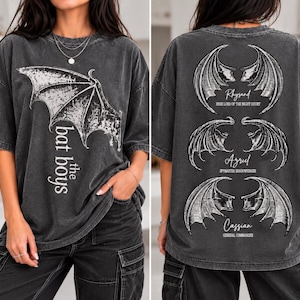 Peut inclure: Un t-shirt gris anthracite à manches courtes avec un motif d'ailes de chauve-souris et le texte "the bat boys" sur le devant. Le dos présente trois paires d'ailes avec les noms "Rhysand", "Azriel" et "Cassian" et leurs titres respectifs.