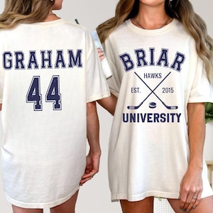 May include: Cream-colored t-shirts with navy blue text. One shirt displays "GRAHAM" above the number "44". The other shirt features "BRIAR HAWKS" with crossed hockey sticks, the text "EST. 2015", and "UNIVERSITY".