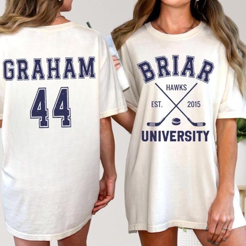 May include: Cream-colored t-shirts with navy blue text. One shirt displays "GRAHAM" above the number "44". The other shirt features "BRIAR HAWKS" with crossed hockey sticks, the text "EST. 2015", and "UNIVERSITY".