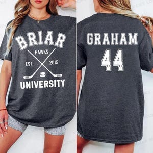 May include: Dark gray t-shirt with white text and graphics. The front features "BRIAR HAWKS UNIVERSITY" with crossed hockey sticks and a puck. The back displays "GRAHAM 44".