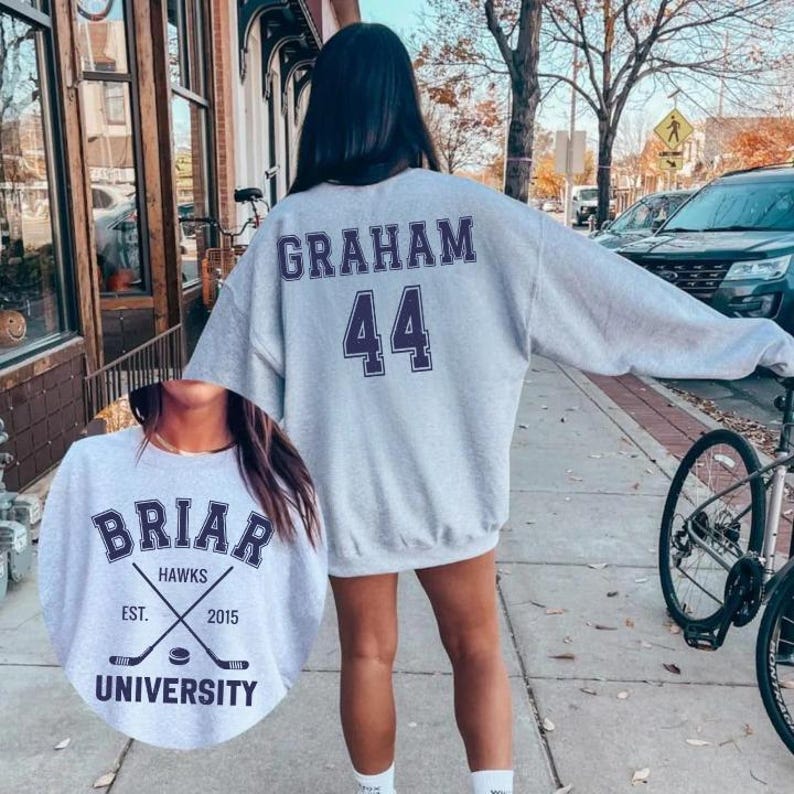 May include: A light gray sweatshirt with the name "GRAHAM" and the number "44" printed on the back in purple. The front of the sweatshirt has a circular graphic with the text "BRIAR HAWKS UNIVERSITY" and crossed hockey sticks.