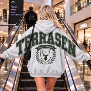 May include: A light grey hooded sweatshirt featuring dark green collegiate-style lettering that reads TERRASEN. Below the text is a dark green crest with a stag head, a star, and laurel branches, with the word GALATHYNIUS on a banner.