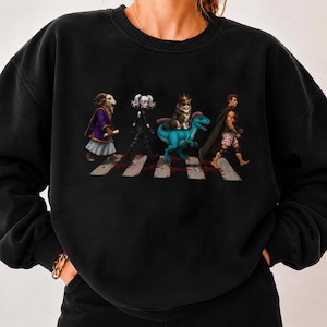 Puede incluir: Una sudadera negra de cuello redondo con un estampado gráfico de cuatro figuras caminando por un paso de cebra. Las figuras incluyen una persona con cabeza de carnero en túnica, una persona con traje negro, un gato con corona sobre un dinosaurio azul y una persona con capa.