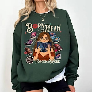 May include: Dark green sweatshirt with the text "Born to Read Forced to Work." The graphic features a woman reading surrounded by books. The design includes red lips, stars, and the phrase "Pages Over Paychecks."
