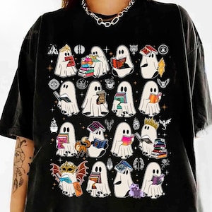 Pode incluir: Uma t-shirt preta com uma impressão gráfica de vários fantasmas brancos a ler livros. Os fantasmas são representados em várias poses, alguns com coroas ou máscaras, rodeados por pequenas ilustrações de estrelas, animais e símbolos místicos.