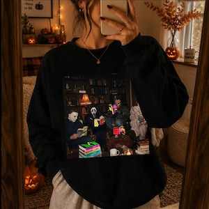 May include: A black long-sleeve sweatshirt featuring a graphic print of four horror movie characters sitting in a library reading books. The print shows figures wearing masks, including a white mask and a ghost mask, surrounded by stacks of books.