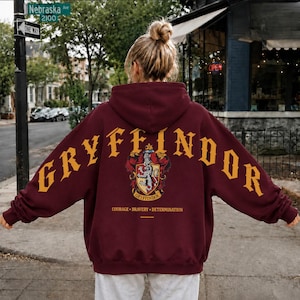 Peut inclure: Un sweat-shirt à capuche bordeaux avec le mot GRYFFINDOR en grandes lettres jaunes arquées dans le dos. Sous le texte se trouve un blason avec un emblème de lion, suivi des mots COURAGE, BRAVERY et DETERMINATION en lettres jaunes.