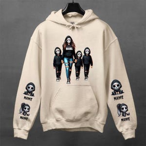 Puede incluir: Sudadera con capucha color crema con un bolsillo frontal grande y una capucha con cordón. La parte delantera presenta un gráfico de una mujer y tres niños con máscaras de hockey. La palabra "MAMA" está impresa encima de la mujer. Las mangas tienen pequeños gráficos con la palabra "NAME".