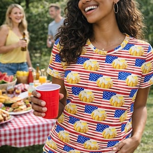 Puede incluir: Camiseta de manga corta con un diseño patriótico con rayas rojas y blancas, banderas azules y pasteles amarillos. La camiseta se combina con pantalones cortos de mezclilla. Se sostiene una taza de plástico roja en la mano.
