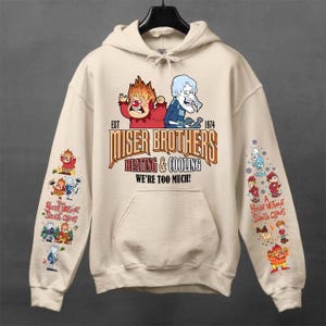 Può includere: Una felpa con cappuccio color crema con una grafica dei fratelli Miser e il testo "EST 1974 MISER BROTHERS HEATING & COOLING WE'RE TOO MUCH!". Le maniche mostrano varie illustrazioni di personaggi e il testo "A Year Without Santa Claus".