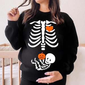 Puede incluir: Una sudadera negra de manga larga con un gráfico blanco de una caja torácica humana y un corazón naranja en el lado izquierdo. Debajo de la caja torácica, hay un gráfico blanco de un pequeño esqueleto sosteniendo una pequeña calabaza naranja.