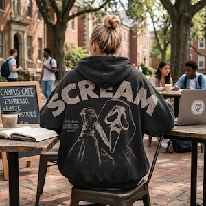 Può includere: Una felpa con cappuccio nera con una grafica invecchiata della maschera di Ghostface dal film Scream sul retro. Il testo recita SCREAM in grandi lettere sopra la maschera e hello there, what's your favorite scary movie? sotto.