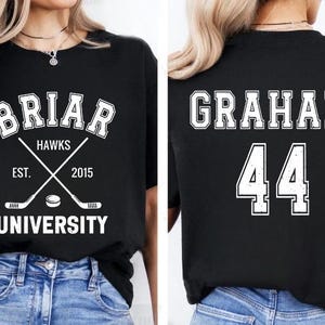May include: Black t-shirt with white text and graphics. The front features "BRIAR HAWKS UNIVERSITY" with crossed hockey sticks and a puck. The back displays "GRAHAM 44". A collegiate-themed apparel item.