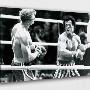 Puede incluir: Impresión en lienzo en blanco y negro de un combate de boxeo. Dos boxeadores musculosos están en el ring, uno con pelo rubio y otro con pelo oscuro, ambos con guantes de boxeo y pantalones cortos. La imagen captura un momento de intensa acción.