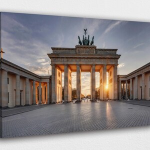 Op de afbeelding: Een canvas print van de Brandenburger Tor in Berlijn, Duitsland, bij zonsondergang. Het neoclassicistische monument heeft hoge zuilen en een quadriga-sculptuur. De lucht is een mix van blauw en oranje, met de zon die achter de poort ondergaat.