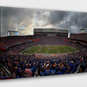 May include: A canvas print of a packed American football stadium during a game. The stands are filled with fans wearing blue and red, and the field is green. The sky is overcast with dark clouds.