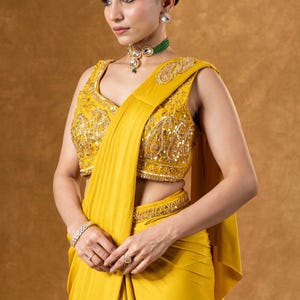 Può includere: Un completo sari giallo senape. Il top senza maniche è impreziosito da paillettes e ricami. Il drappeggio del sari è liscio e fluido. Una collana girocollo di perline verdi e orecchini abbinati completano il look. Sono visibili anche un braccialetto e un anello d'oro.