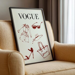 K&ouml;nnte beinhalten: Ein gerahmter Druck mit dem Wort "VOGUE" &uuml;ber roten Skizzen von Modeaccessoires. Die Skizzen umfassen einen Mantel, eine Sonnenbrille, einen Hut, eine Handtasche und High-Heels. Der Druck wird auf einem beigen Sessel pr&auml;sentiert.