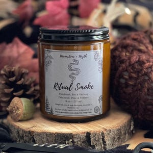 May include: A glass jar candle with a black lid, labeled "Ritual Smoke." The label includes the text "Moonstone Myth" and lists ingredients. The candle sits on a wooden slice, with a pair of black candle scissors in the foreground. A knitted pumpkin and fall foliage are in the background.