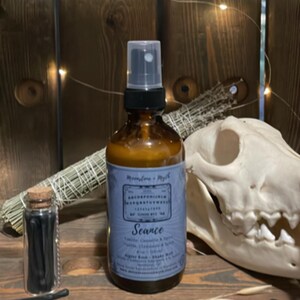 Peut inclure: Une bouteille de pulvérisation en verre brun avec une étiquette bleue indiquant "Seance" et "Moonstone + Myth". À côté, un petit pot en verre avec un bouchon, un bouquet d'herbes séchées et un crâne de chien. Le fond est une surface en bois.