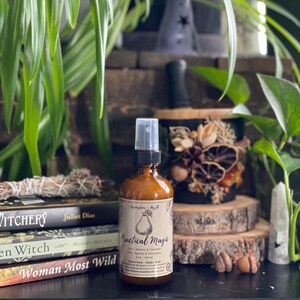 Peut inclure: Un flacon pulvérisateur en verre ambré avec un embout noir et une étiquette beige illustrée d'une poire et du texte Practical Magic, Pear, Apricot & Cinnamon, 118 ml. Le flacon est placé devant des livres empilés et un décor naturel.