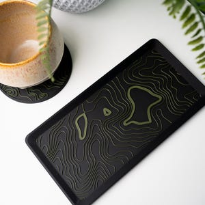 May include: A black rectangular tray with an olive green topographic map design. A matching coaster with the same design is visible. The tray is approximately 30 cm long and 20 cm wide.