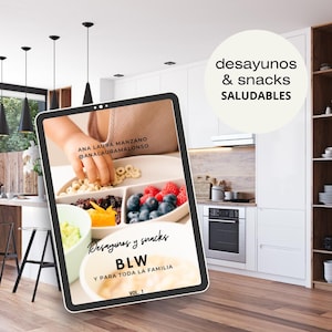 Puede incluir: Una tableta muestra un desayuno colorido con bayas, cereales y frutas. El texto en la tableta dice "Desayunos y snacks BLW Y PARA TODA LA FAMILIA". El fondo muestra una cocina moderna con gabinetes blancos y luces colgantes negras.