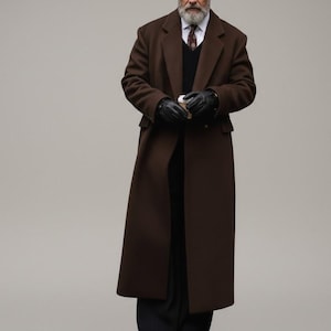 Puede incluir: Un abrigo largo marrón con un diseño clásico. El abrigo presenta una solapa con muescas, un cierre cruzado y un ajuste a medida. La persona lleva un suéter negro, una camisa blanca y una corbata estampada. Se usan guantes negros.
