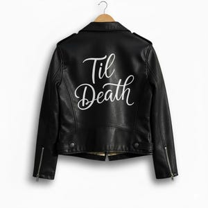 以下が含まれることがあります： 背中に白い筆記体で「Til Death」と書かれた黒いフェイクレザーのジャケット。ノッチカラー、非対称ジッパー、ジッパーポケットを備えたクラシックなバイカースタイルです。木製のハンガーにかかっています。
