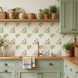 Può includere: Un paraschizzi da cucina con piastrelle bianche quadrate decorate con motivi floreali dipinti a mano in blu e giallo. La cucina presenta armadietti color verde salvia, un piano di lavoro in legno, un rubinetto in rame e un asciugamano.