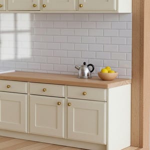 May include: A miniature kitchen scene featuring off-white cabinets with brass knobs, a light wood countertop, and a white subway tile backsplash. A small silver metal teapot and a wooden bowl containing three yellow lemons sit on the counter.