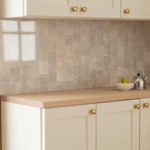 May include: Off-white kitchen cabinets with brass knobs, a light wood countertop, and a backsplash featuring rectangular tiles in varying shades of beige and gray. A small white bowl holds green limes next to a silver metal tea kettle.