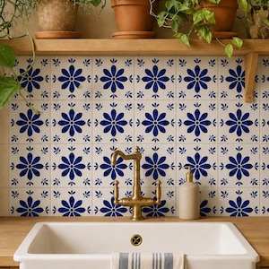 Op de afbeelding: Een witte boerderijspoelbak met een messing kraan en een gestreepte handdoek, voor een achterwand van witte vierkante tegels met een blauw bloemmotief. Boven de spoelbak staat een houten plank met diverse groene planten in potten.