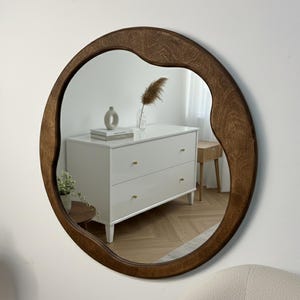 Puede incluir: Un espejo de pared redondo con un marco de madera grueso de color marrón oscuro con un borde irregular y ondulado. La superficie reflectante muestra el interior de una habitación con una cómoda blanca, una planta pequeña y un jarrón decorativo.