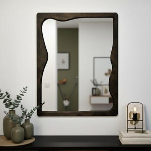 May include: A dark brown, uniquely shaped mirror hangs on a white wall. The mirror's frame has an organic, wavy design. Below, a black table holds vases with greenery and a decorative lamp with a lightbulb.
