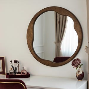 Peut inclure: Un miroir rond encadré de bois de forme organique est suspendu au-dessus d'une coiffeuse blanche. Un plateau bordeaux contient des pinceaux de maquillage et des flacons de parfum. Une photo encadrée et un vase avec une fleur sont également sur la coiffeuse. Des rideaux bordeaux encadrent la fenêtre.