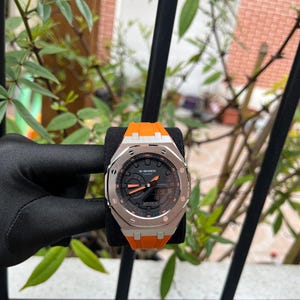 Könnte beinhalten: Eine G-Shock Casio Armbanduhr mit einer silberfarbenen achteckigen Lünette mit sichtbaren Schrauben, einem schwarz-analogen Digitalzifferblatt mit orangefarbenen Akzenten und einem leuchtend orangefarbenen Silikonarmband. Das Zifferblatt zeigt den Markennamen G-Shock Casio.