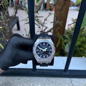 Könnte beinhalten: Eine silberfarbene G-Shock-Uhr mit schwarzem Zifferblatt und schwarzem Kautschukarmband. Die Uhr zeigt Datum und Uhrzeit digital an. Die Uhr wird auf einer schwarzen Oberfläche präsentiert.