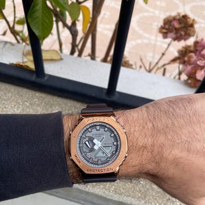 Könnte beinhalten: Eine G-Shock Digitaluhr mit braunem Armband und goldfarbener Lünette. Das Zifferblatt ist grau mit digitaler Anzeige und analogen Zeigern. Die Uhr befindet sich an einem Handgelenk.