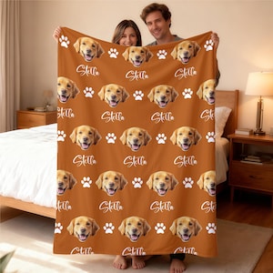 Może przedstawiać: Spersonalizowany pomarańczowy koc z powtarzającym się wzorem twarzy golden retrievera, białymi odciskami łap i imieniem "Stella" w ozdobnej czcionce. Koc jest trzymany przez dwie osoby.