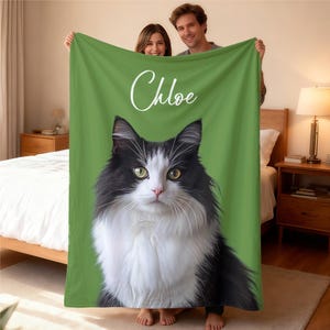 May include: A green blanket featuring a black and white cat with yellow eyes and the name "Chloe" in white script. The cat's face is centered on the blanket, with a green background. The blanket is held up by two people.