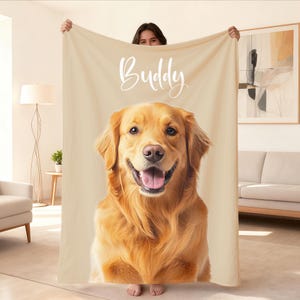 May include: A beige blanket featuring a golden retriever dog with the name "Buddy" in white script. The dog has a happy expression with its tongue out. The blanket is held up by a person.