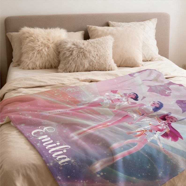 May include: A personalized blanket with a soft, colorful design featuring three figures in a flowing, ethereal style. The blanket is predominantly pink and purple, with the name "Emilia" printed in white script. The blanket is on a bed with fluffy pillows.