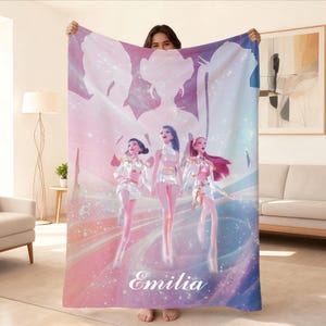 May include: A large, rectangular blanket featuring a digital illustration of three stylized female figures in white and gold outfits. The blanket has a soft color palette of pink, blue, and purple, with the name "Emilia" printed in white.