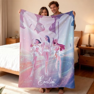 May include: A soft blanket with a colourful design of three figures in white and silver outfits, set against a pastel background. The name "Emilia" is written in elegant script across the bottom of the blanket.