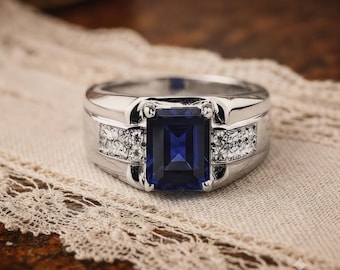 May include: A silver-toned ring featuring a large, rectangular, emerald-cut deep blue gemstone set in the center. The band displays intricate, textured floral-like engravings on both sides of the central stone setting, creating a detailed finish.
