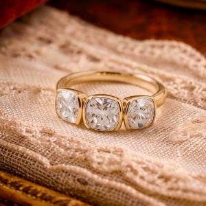May include: A gold ring featuring three cushion-cut clear stones set in a row. The band is smooth and polished, holding the stones in individual bezel settings. The design showcases a central larger stone flanked by two smaller stones of the same shape.