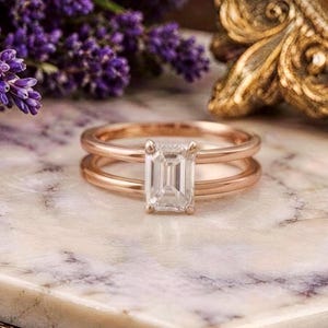 May include: A rose gold ring featuring a clear emerald-cut gemstone set in a four-prong setting. The band consists of two parallel thin rose gold wires that meet at the centre to hold the rectangular stone securely in place.